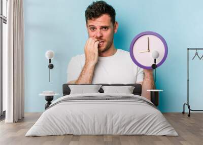 Young caucasian man holding a clock isolated on blue background biting fingernails, nervous and very anxious. Wall mural