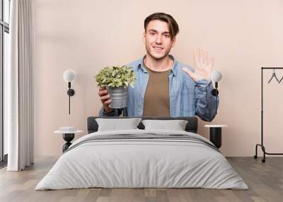 Young caucasian man holding a plant smiling cheerful showing number five with fingers. Wall mural