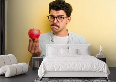 Young caucasian man holding an apple isolated on yellow background confused, feels doubtful and unsure. Wall mural