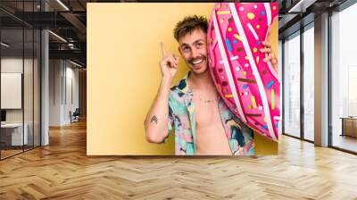 Young caucasian man holding an inflatable donut isolated on yellow background Wall mural