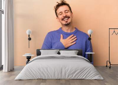 Young caucasian man isolated on beige background laughs out loudly keeping hand on chest. Wall mural