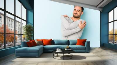 Young caucasian man isolated on blue background hugs, smiling carefree and happy. Wall mural