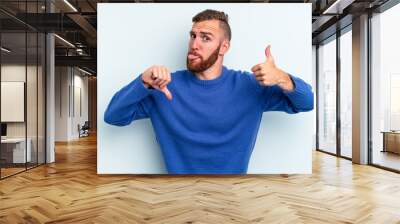 Young caucasian man isolated on blue background showing thumbs up and thumbs down, difficult choose concept Wall mural