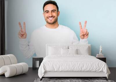 Young caucasian man isolated on blue background showing victory sign and smiling broadly. Wall mural