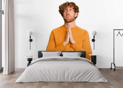 Young caucasian man isolated on green chroma background holding hands in pray near mouth, feels confident. Wall mural