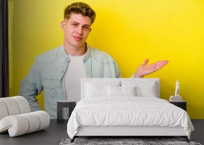 Young caucasian man isolated on yellow background showing a copy space on a palm and holding another hand on waist. Wall mural