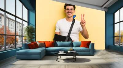 Young caucasian man with broke hand isolated on yellow background joyful and carefree showing a peace symbol with fingers. Wall mural