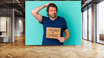 Young caucasian middle age man holding a We need a change placard isolated on blue background Wall mural