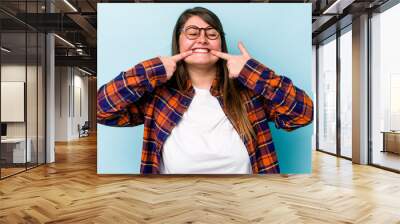 Young caucasian overweight woman isolated on blue background smiles, pointing fingers at mouth. Wall mural