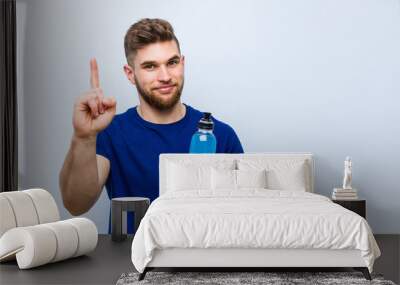 Young caucasian sportsman holding an isotonic drink showing number one with finger. Wall mural