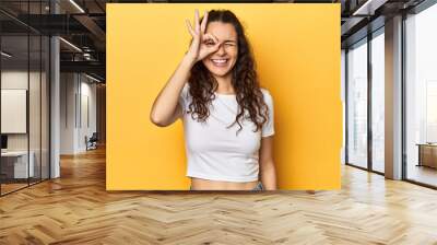 Young Caucasian woman, yellow studio background, excited keeping ok gesture on eye. Wall mural