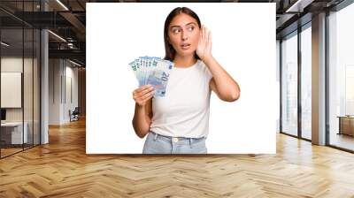 Young caucasian woman holding a banknotes isolated trying to listening a gossip. Wall mural