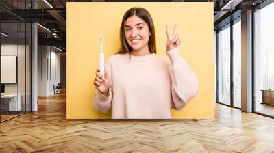 Young caucasian woman holding a electric toothbrush isolated on yellow background showing number two with fingers. Wall mural