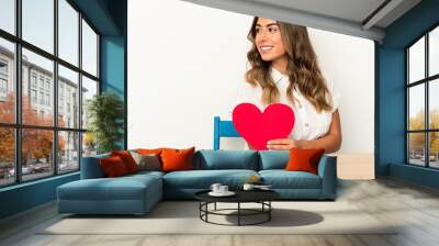 Young caucasian woman holding a valentines day heart isolated looks aside smiling, cheerful and pleasant. Wall mural