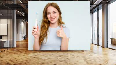 Young caucasian woman holding electric toothbrush isolated on blue background smiling and raising thumb up Wall mural