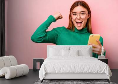 Young caucasian woman holding mobile phone isolated on pink background raising fist after a victory, winner concept. Wall mural