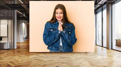 young caucasian woman isolated en beige background holding hands in pray near mouth, feels confident Wall mural