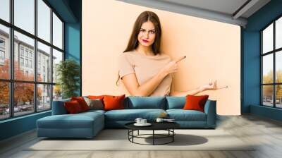 Young caucasian woman isolated en beige background shocked pointing with index fingers to a copy space. Wall mural