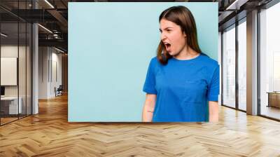 Young caucasian woman isolated on blue background shouting very angry, rage concept, frustrated. Wall mural