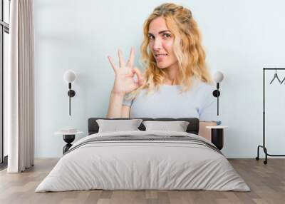 Young caucasian woman isolated on blue background winks an eye and holds an okay gesture with hand. Wall mural