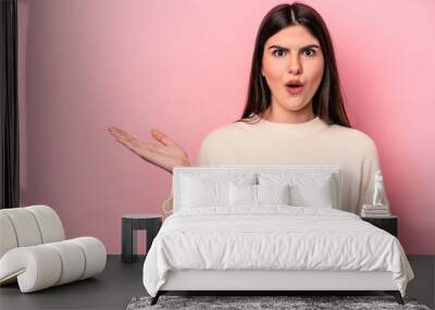 Young caucasian woman isolated on pink background impressed holding copy space on palm. Wall mural