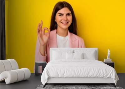 Young caucasian woman isolated on yellow background winks an eye and holds an okay gesture with hand. Wall mural