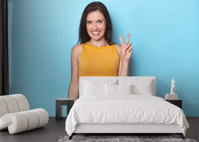 Young caucasian woman on blue backdrop showing victory sign and smiling broadly. Wall mural