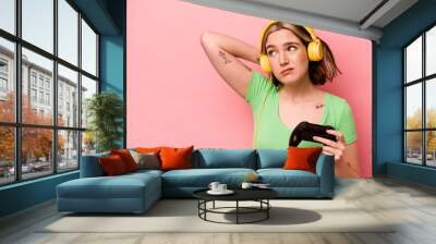 Young caucasian woman playing with a video game controller isolated on pink background touching back of head, thinking and making a choice. Wall mural