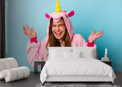 Young caucasian woman wearing a unicorn pajama isolated on blue background receiving a pleasant surprise, excited and raising hands. Wall mural