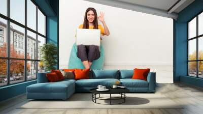 Young caucasian woman working with laptop sitting on a puff isolated on white background cheerful and confident showing ok gesture. Wall mural