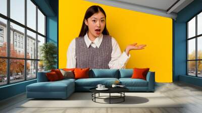 young chinese woman isolated on yellow background impressed holding copy space on palm. Wall mural