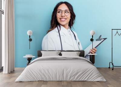 Young doctor mexican woman isolated on blue background looks aside smiling, cheerful and pleasant. Wall mural