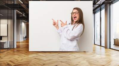 Young doctor woman against a white wall pointing with forefingers to a copy space, expressing excitement and desire. Wall mural