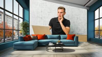 Young entrepreneur working with his laptop on a desk is saying a secret hot braking news and looking aside Wall mural