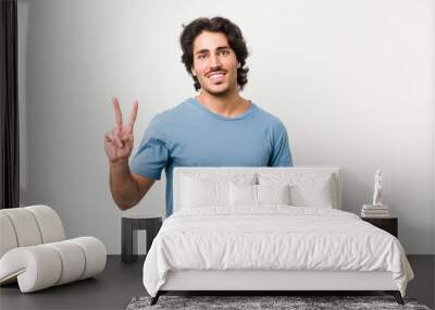 Young handsome man against a white background showing victory sign and smiling broadly. Wall mural