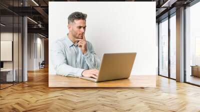 Young handsome man working with his laptop looking sideways with doubtful and skeptical expression. Wall mural