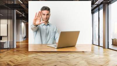 Young handsome man working with his laptop standing with outstretched hand showing stop sign, preventing you. Wall mural