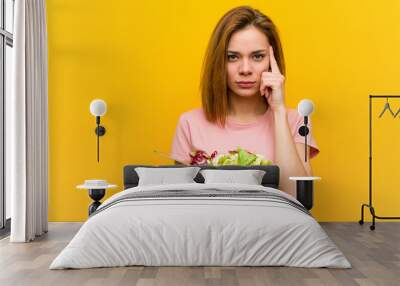 Young healthy woman holding a salad pointing her temple with finger, thinking, focused on a task. Wall mural