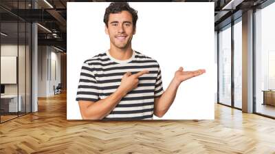 Young Hispanic man in studio excited holding a copy space on palm. Wall mural