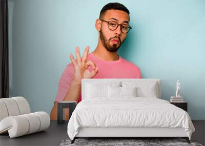 Young hispanic man isolated on blue background winks an eye and holds an okay gesture with hand. Wall mural