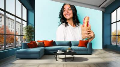 Young hispanic woman eating a sandwich isolated on blue background looks aside smiling, cheerful and pleasant. Wall mural