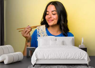 Young hispanic woman eating noodles isolated on yellow background Wall mural