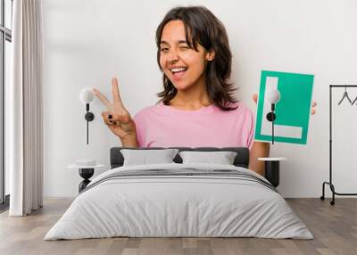 Young hispanic woman holding a beginner driver sign isolated on white background joyful and carefree showing a peace symbol with fingers. Wall mural