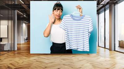 Young hispanic woman holding a t-shirt on a hanger isolated on blue background trying to listening a gossip. Wall mural