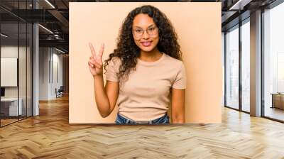 Young hispanic woman isolated on beige background showing victory sign and smiling broadly. Wall mural