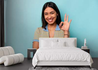 Young hispanic woman isolated on blue background winks an eye and holds an okay gesture with hand. Wall mural