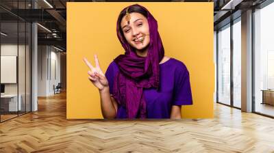 Young Indian woman wearing a traditional sari clothes isolated on yellow background joyful and carefree showing a peace symbol with fingers. Wall mural