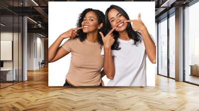 Young Latin friends in studio smiles, pointing fingers at mouth. Wall mural