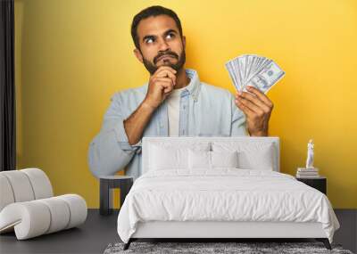 Young Latino man holding a bundle of dollars, yellow studio background, looking sideways with doubtful and skeptical expression. Wall mural