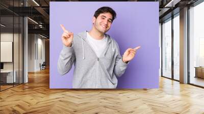 Young man isolated on purple background pointing to different copy spaces, choosing one of them, showing with finger. Wall mural